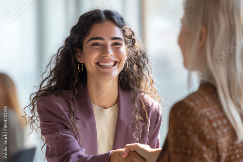 Young woman with curly hair smiling warmly while shaking hands with another person in a professional setting, symbolizing agreement or a positive interaction