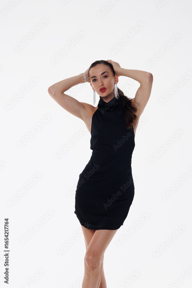 Fototapeta premium Beautiful young woman in a black elegant dress posing in studio on white background. Fashion model with long hair, red lips, and stylish earrings. Glamour portrait, studio photoshoot, beauty 