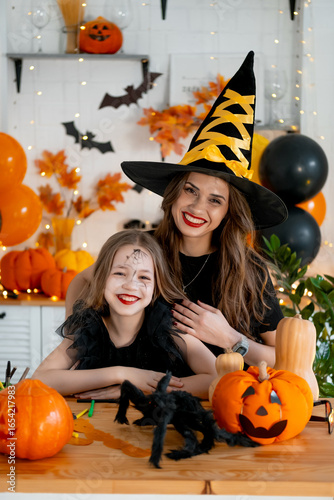 A happy family is getting ready for Halloween. Mother and child daughter in carnival costumes at home in the kitchen having fun scaring each other and laughing
