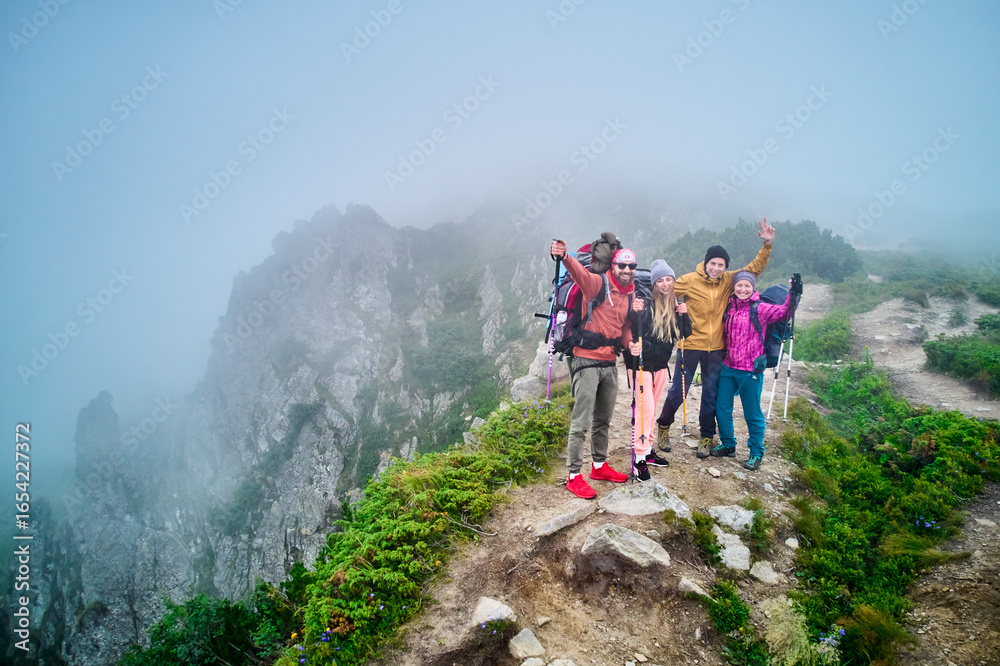 Obraz premium Group of four hikers, equipped with backpacks and trekking poles, cheerfully pose on misty mountain ridge. Rocky cliffs, lush greenery around create dramatic backdrop, while fog adds mystical touch.