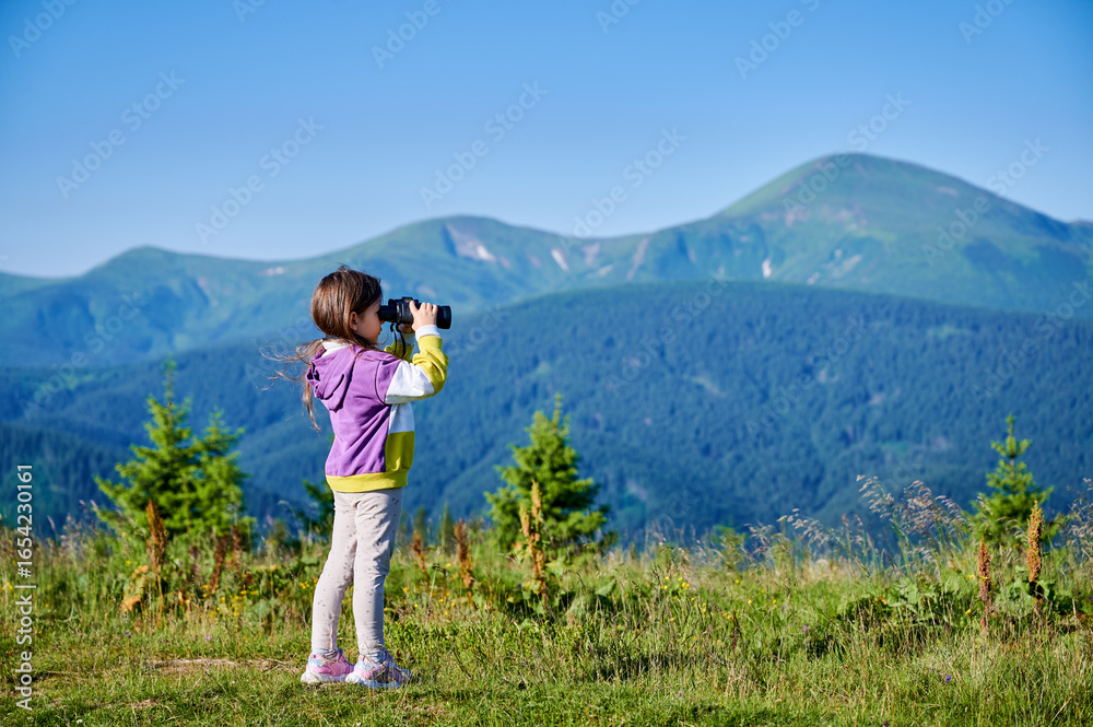 Naklejka premium Young girl stands in grassy meadow, looking through binoculars, wearing yellow and purple sweatshirt. Behind her, rolling green hills and mountains stretch into distance under clear blue sky.