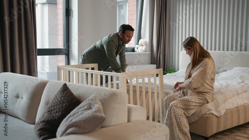 A pregnant woman sits on the bed and looks for details from the crib while a man stands next to her