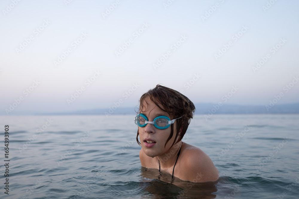 Naklejka premium Boy resting in swimming goggles, floating peacefully on calm lake waters during golden sunset, nature embracing swimmer's tranquil moment
