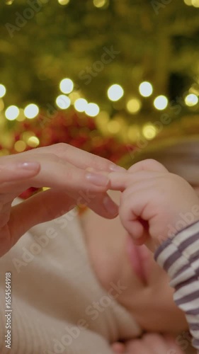 Vertical Screen: A young mom lies on a cozy blanket next to the Christmas tree, breastfeeding her baby in a warm holiday setting filled with lights and decorations.