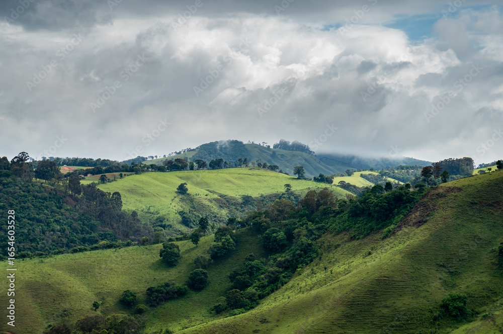 Obraz premium Dramatic cloudy sky over rolling green hills and valleys of Monte Verde in Mantiqueira mountains, with scattered trees and lush vegetation patches in Camanducaia, Minas Gerais - Brazil