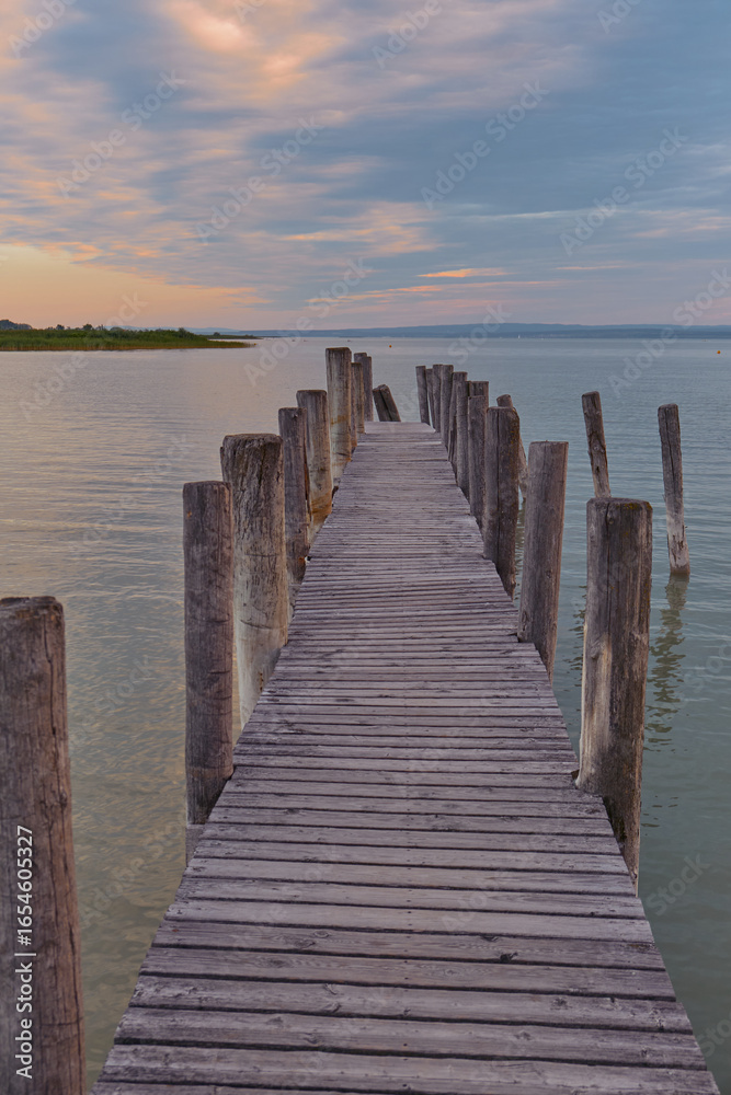 Fototapeta premium Atmospheric wooden pier stretching into a calm lake with vibrant sunset sky reflecting soft golden and peach tones, creating a mysterious and tranquil mood