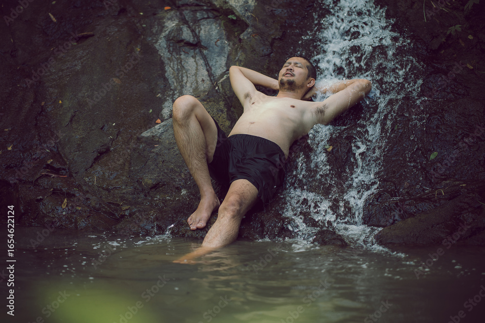 Fototapeta premium A Young Asian man in lotus yoga pose meditating on a peaceful natural waterfall. Beautiful natural sculpture of art and religion.