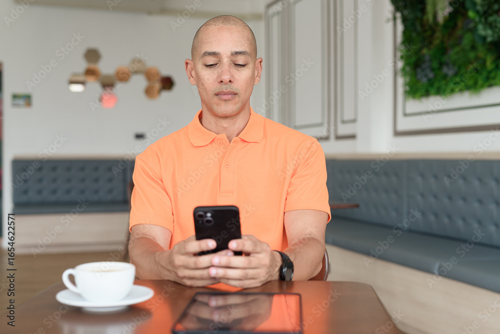 Bald Hispanic man with coffee using phone sending sms in cafe restaurant wearing orange polo shirt Bald Hispanic man with coffee using phone sending sms in cafe restaurant wearing orange polo shirt