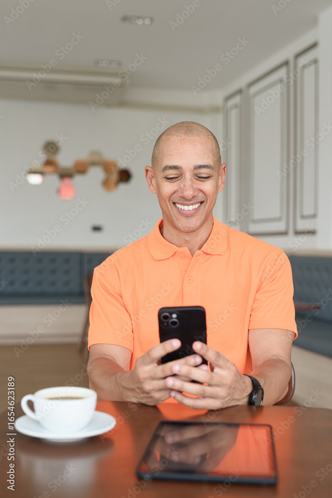 Bald Hispanic man with coffee using phone smiling in restaurant wearing orange polo shirt Bald Hispanic man with coffee using phone smiling in restaurant wearing orange polo shirt