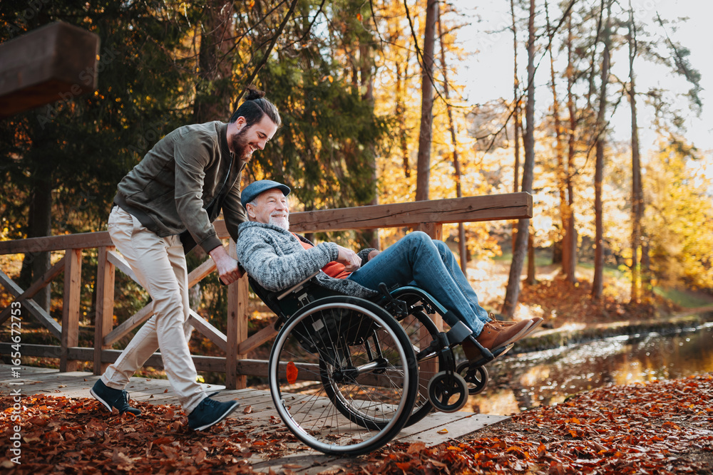 Grandson pushing his older grandad in wheelchair during walk in fall nature. Grandson pushing his older grandad in wheelchair during walk in fall nature.