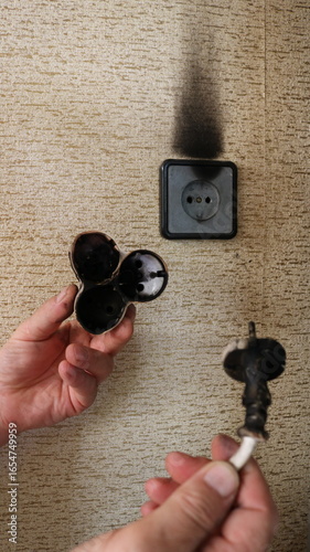 male hands holding scorched wire and tee against background of wall home socket with black traces of burning, dangerous domestic incident related to risk of fire due to malfunction of electrical