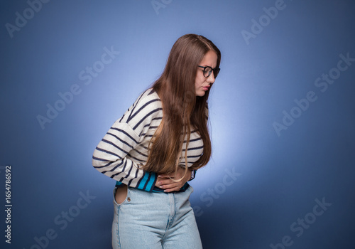 Young woman suffering from bellyache - stomachache, isolated on blue background. Ill girl with gastritis. Abdomen bloating concept.