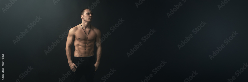 Fototapeta premium Muscular Man with Rosary on Dark Wide Banner with Copy Space