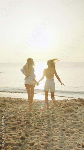 Two female friends joyfully running along the beach at sunset, creating lasting memories on a warm summer evening,vertical video