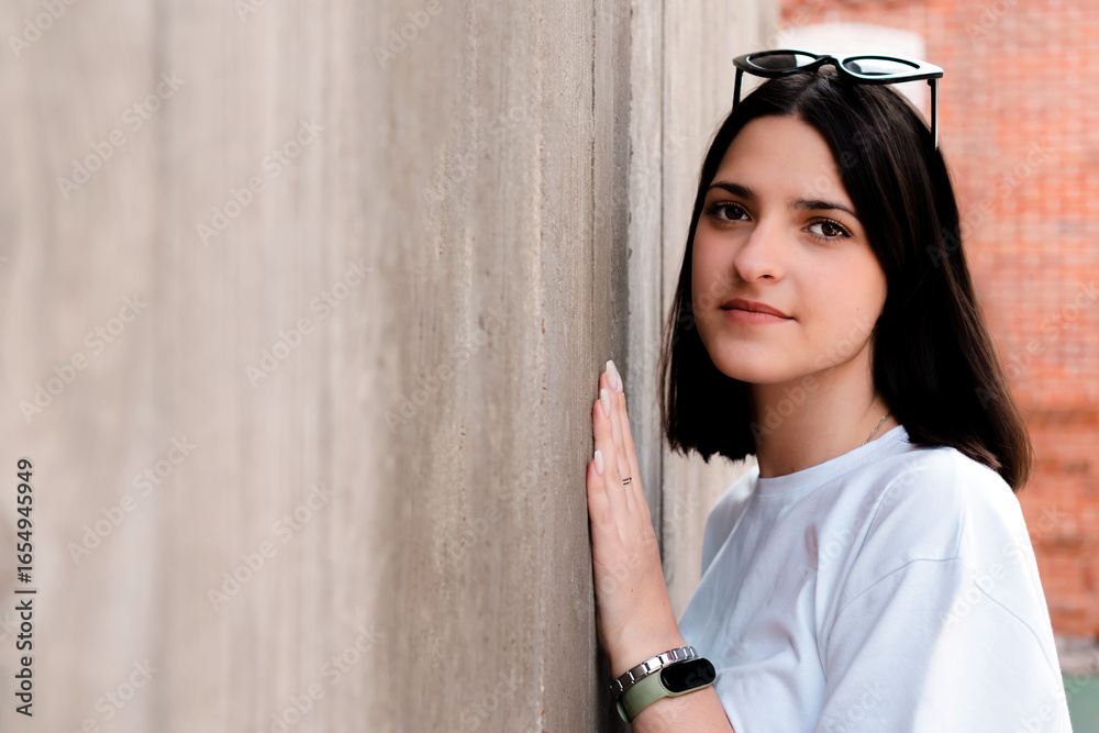 Obraz premium Young woman with sunglasses on her head and smartwatch on her wrist gently touching a concrete wall with her hand, creating a sense of casual coolness and urban style