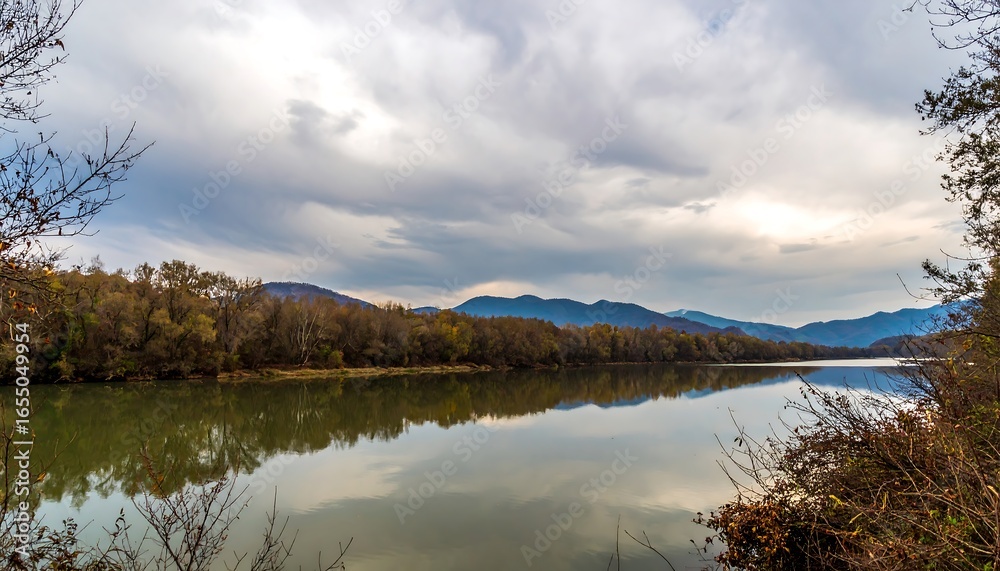 Fototapeta premium Tranquil autumnal scene of a calm river reflecting a cloudy sky and forested hills.