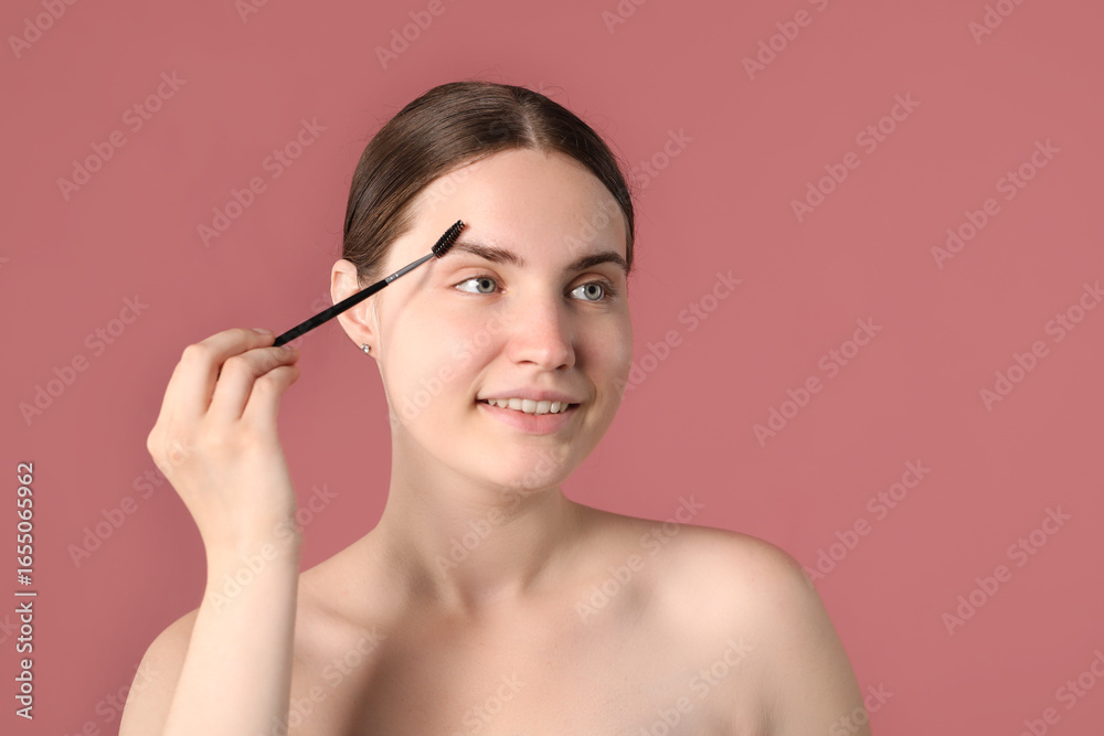 Fototapeta premium Young woman brushing her eyebrow with spoolie on pink background
