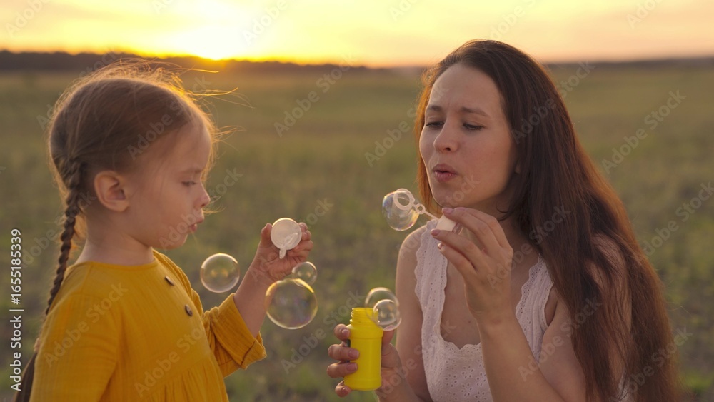 Fototapeta premium Happy Mother and her little Daughter playing together outdoor, blowing soap bubbles, having fun on backyard. Nature. Beauty Mum and her Child in Park together. Family, Mother's Day Joy. Mom and Baby