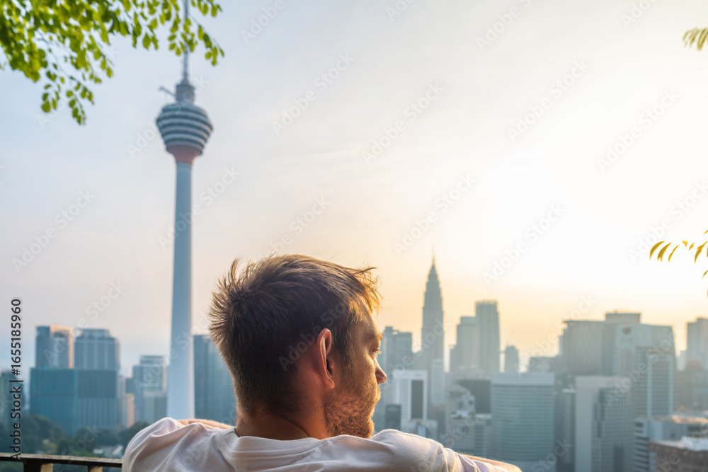 Obraz premium Thoughtful man standing on rooftop terrace with green plants and looking at panoramic view of Kuala Lumpur city with tower at sunrise, Malaysia. Sleepy male tourist looking at skyline of Asian city