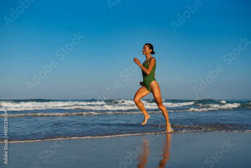 Cheerful woman in green swimsuit running along the beach in shallow water, enjoying the sunlight and ocean waves on a bright summer morning, smiling with freedom and energy.