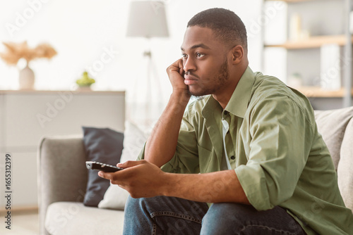 Tableau sur toile Bored African American Man Watching Boring TV Switching Channels Holding Television Remote Controller Sitting On Sofa At Home