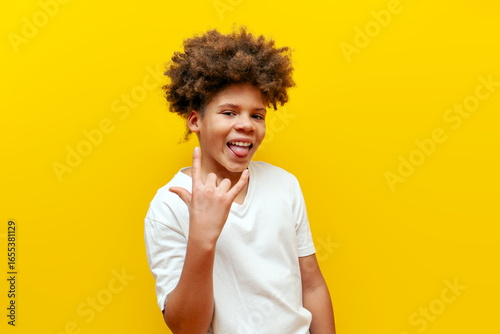 Valokuva cheerful curly african american boy showing rock music gesture and tongue on yel