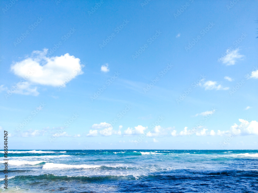 Obraz premium The surface of the sea water ripples with the horizon line meeting the blue sky and wisps of white clouds