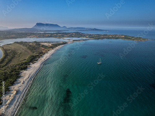 Aerial view of a lone sailboat glides on the turquoise waters near the pristine white sands of La Cinta Beach, backed by the majestic Tavolara Island, San Teodoro, Sardinia, Italy.
