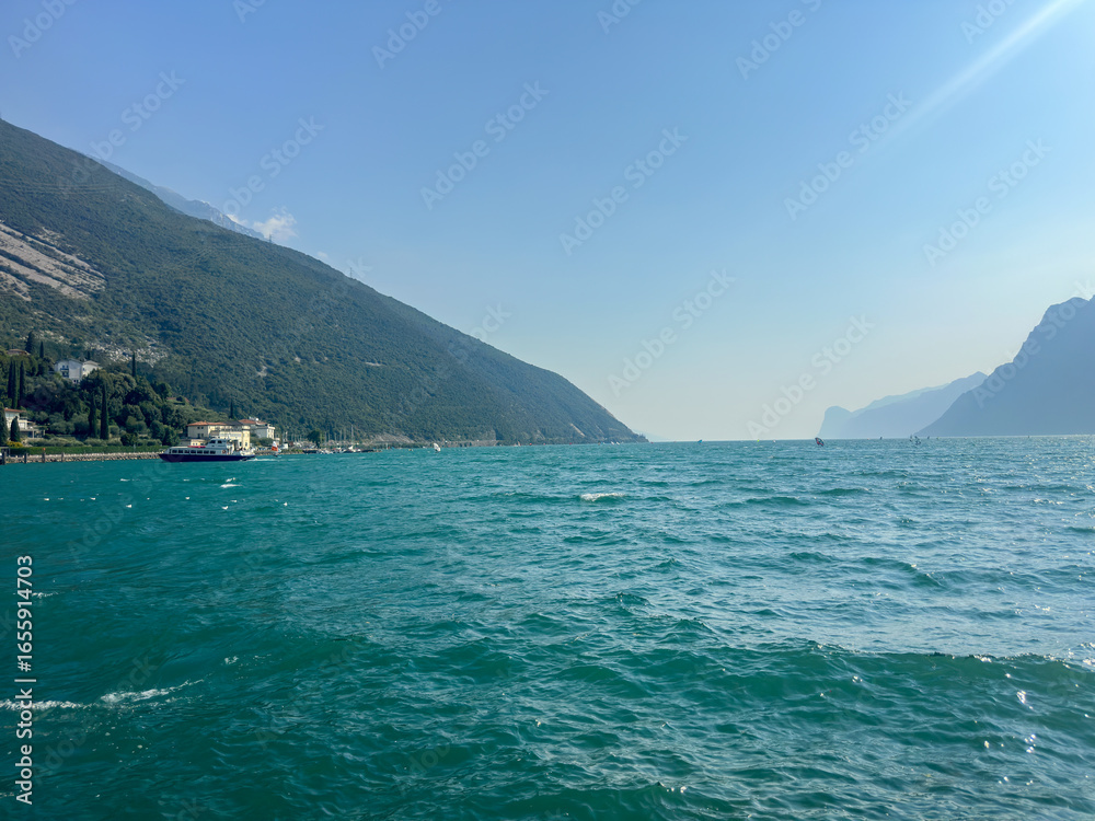 Fototapeta premium Scenic view of the turquoise waters of Lake Garda from a rocky promenade in Torbole, with mountains and buildings in the background. High quality photo