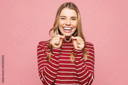 Young smiling satisfied woman wearing red striped sweatshirt casual clothes hold in hand invisible transparent aligners, invisalign dental braces isolated on plain pink background. Lifestyle concept.