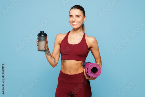 фотографія Young fitness trainer instructor woman sportsman wear red sportswear training in home gym hold yoga mat drink protein shake cocktail isolated on plain blue background