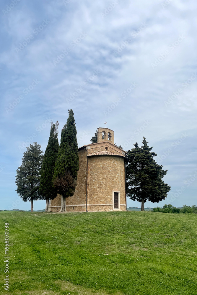 Fototapeta premium Small stone chapel surrounded by cypress trees on a grassy hill in the Tuscan countryside under a partly cloudy spring sky