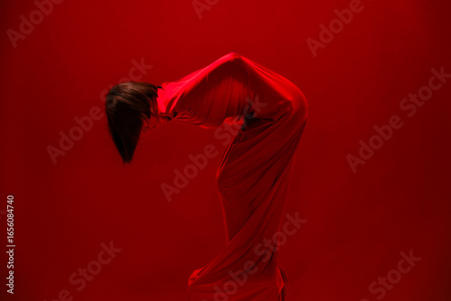 A person is fully covered by a flowing red fabric, bending deeply with their head bowed. Dark hair cascades downwards. The monochromatic red setting emphasizes the dramatic and mysterious pose.