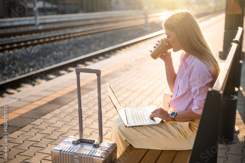 Elegant businesswoman using laptop and drinking coffee while sitting on a bench with suitcase at the  railway station and waiting arrival of train