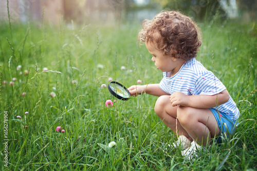 beautiful curly little toddler girl exploring nature with magnifying glass in summer,  Child natural science education concept