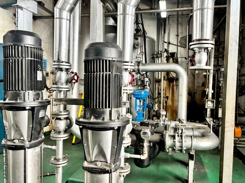 Industrial water pumping station. Close-up of vertical centrifugal pumps driven by electric motors, connected to a stainless steel piping system in a modern factory, utility, or HVAC room, representin