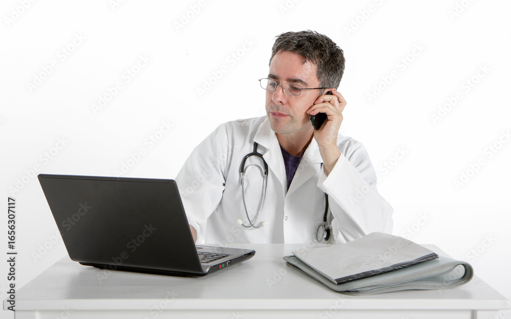 Medical Admin: Second Opinion. A doctor uses his laptop while on a phone call sitting at his desk with a laptop, a notebook, and a stethoscope. Medical Admin: Second Opinion. A doctor uses his laptop while on a phone call sitting at his desk with a laptop, a notebook, and a stethoscope.
