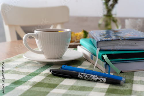 Cozy morning scene with white cup of coffee on saucer, stack of colorful notebooks, pens, and green checkered tablecloth on wooden table, blurred background with chair and vase. Planning and reading .