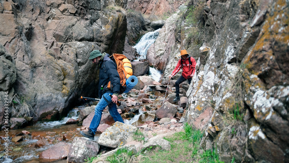 Two active men are having a great time hiking in the mountains. The boys go down a rocky path, next to which a stream flows Two active men are having a great time hiking in the mountains. The boys go down a rocky path, next to which a stream flows