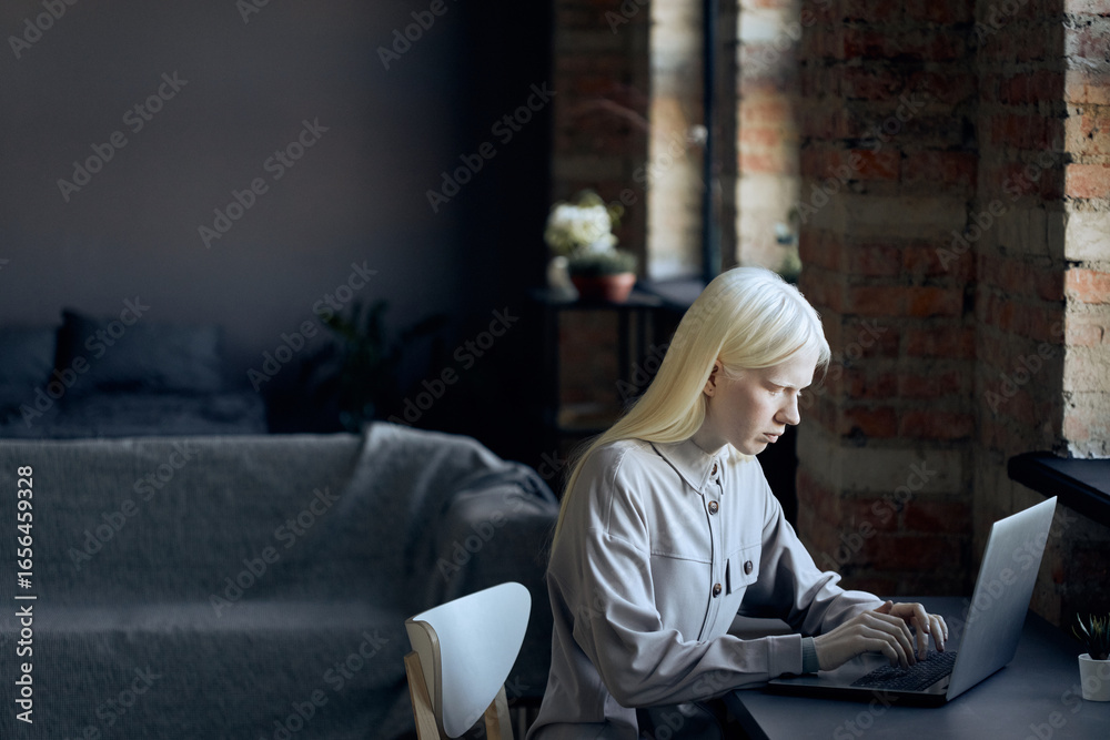 Obraz premium Androgynous young woman with albinism sitting at desk in loft living room at home surfing Internet on laptop