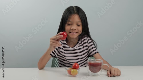 A cute kid girl wearing a patterned shirt picks up a red tomato from the bowl to show, while holding a glass of fresh tomato juice in the other hand to drink and raising her thumb with gratitude.