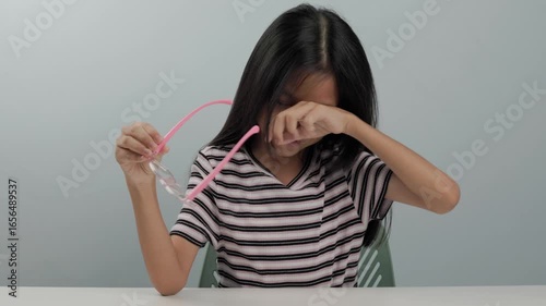 A kid primary girl is wearing glasses to block blue light from a screen. She removes her glasses and rubs her left eye because it feels irritated, itchy, and uncomfortable. The background is plain.