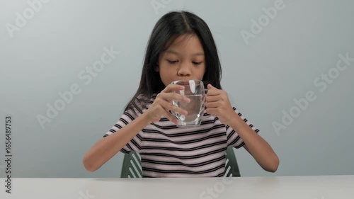 A cute girl wearing a striped shirt with long black hair lifted a glass of water on a white table and took a sip to quench her thirst. When your throat is parched, drinking water is refreshing.