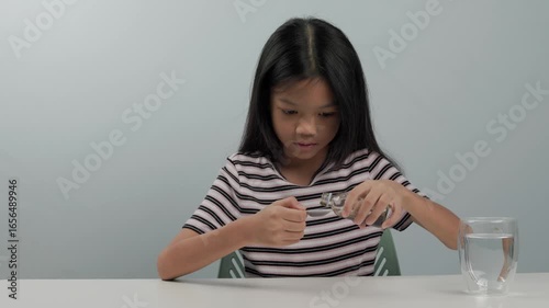 A cute child girl wearing a striped shirt and long black hair is carefully pouring black liquid medicine from a small medicine bottle into two full white teaspoons and drinking them by herself.