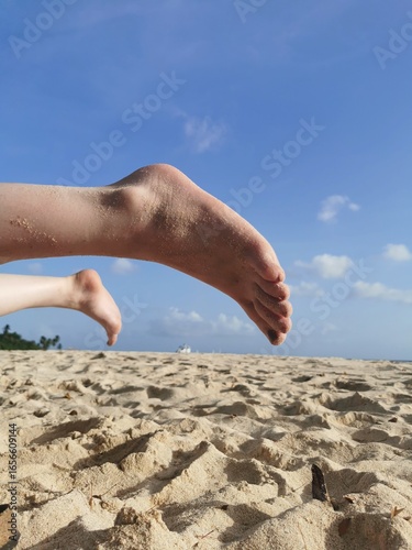 Bare feet playfully extended over pristine, sun-kissed sand with a blue sky and ocean horizon. Perfect for travel, relaxation, and vacation themes.
