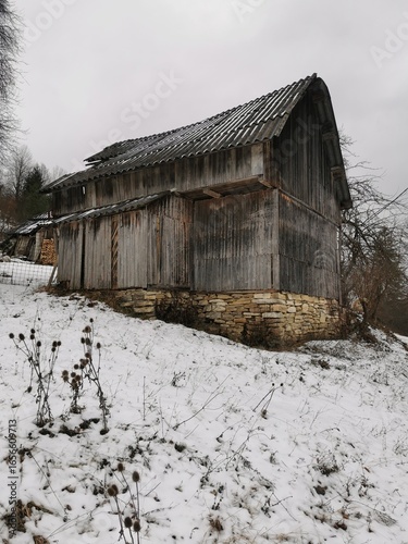 A weathered wooden barn on a hill with a stone foundation, surrounded by snow.