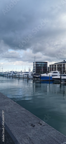 A row of expensive yachts and boats docked in a modern marina on a cloudy day. The water reflects the buildings and dramatic sky.