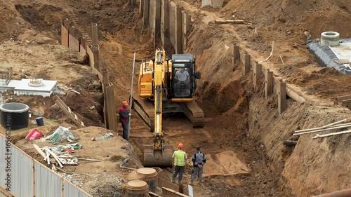 An overhead shot of a large, deep trench at a construction site, where an excavator and several workers are performing excavation work