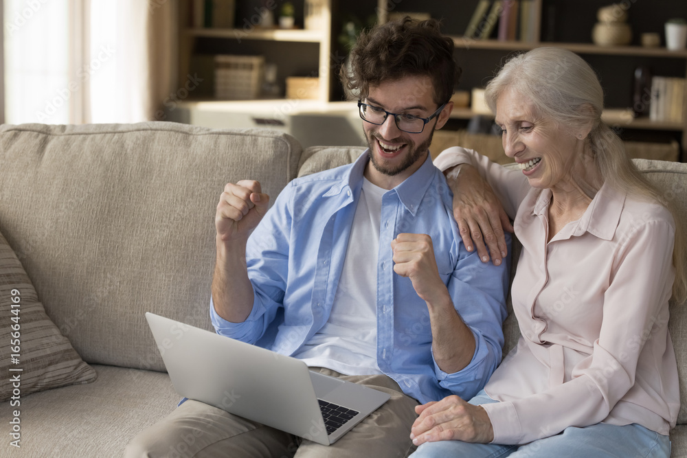 Fototapeta premium We did it. Overjoyed adult son hold notebook on lap raise hands up in victory gesture celebrate online success together with senior mother winning internet game getting bonus lottery prize. Copy space