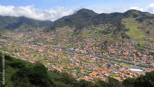 Machico Madeira, Portugal. A town with many houses featuring red-tiled roofs is spread across a wide valley, surrounded by tall green mountains partly covered in clouds.